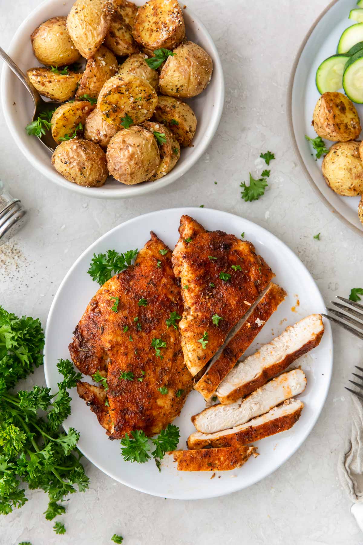 Two air fryer chicken breasts partially sliced on a plate with a bowl of potatoes in the background.