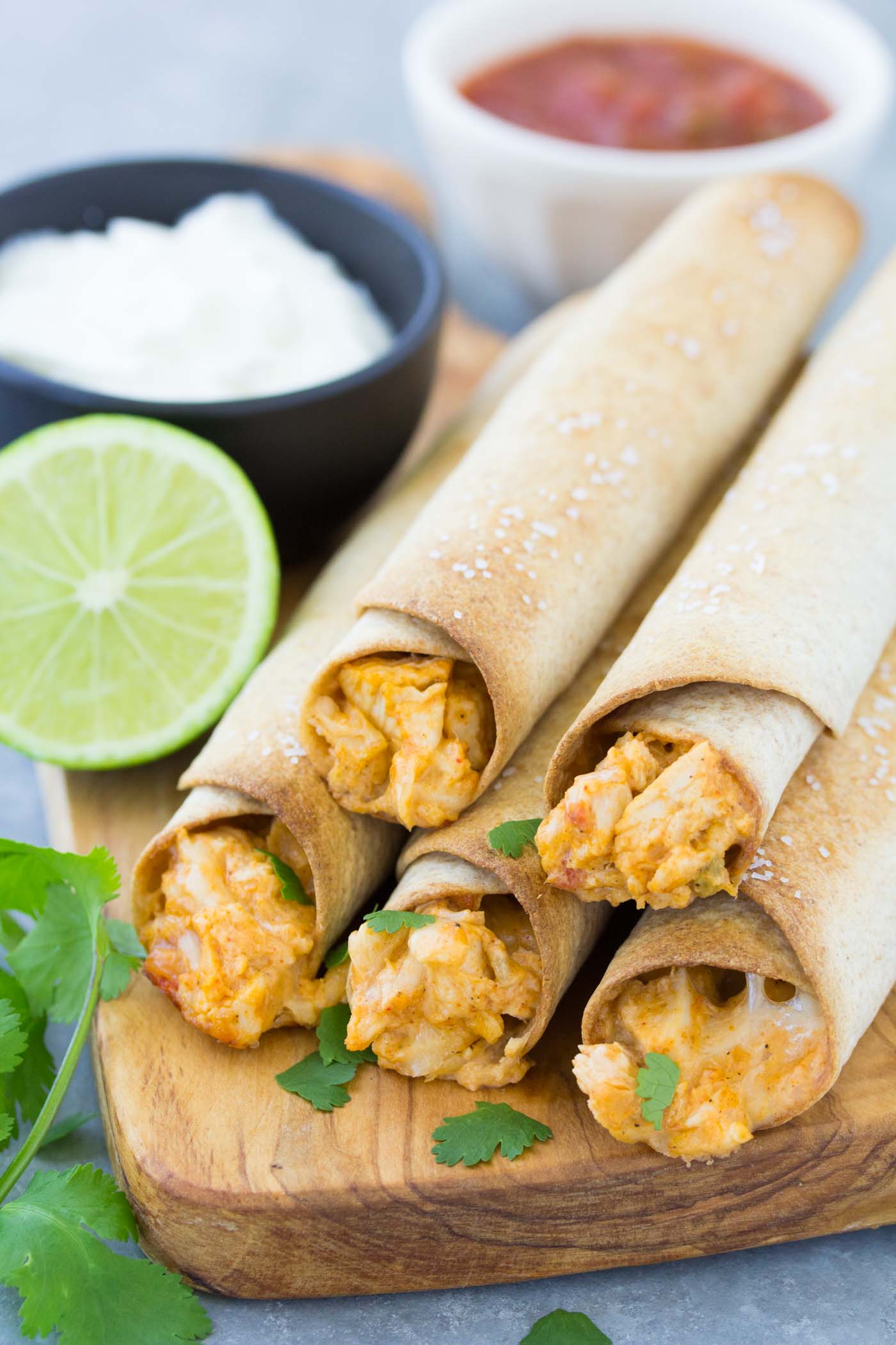 Five baked chicken taquitos stacked together with dishes of plain Greek yogurt and salsa in the background.