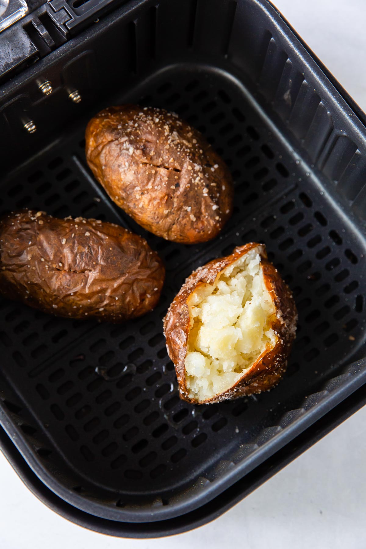 Three baked potatoes in air fryer with one potato cut open.