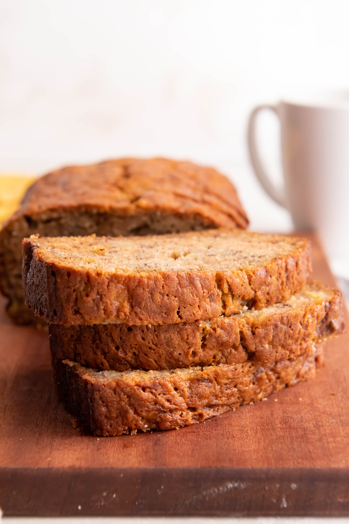 Three slices of banana bread stacked on top of each other with rest of loaf and white mug in background.