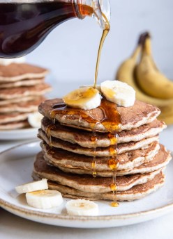 Pouring maple syrup onto a stack of 7 banana pancakes garnished with fresh banana slices.