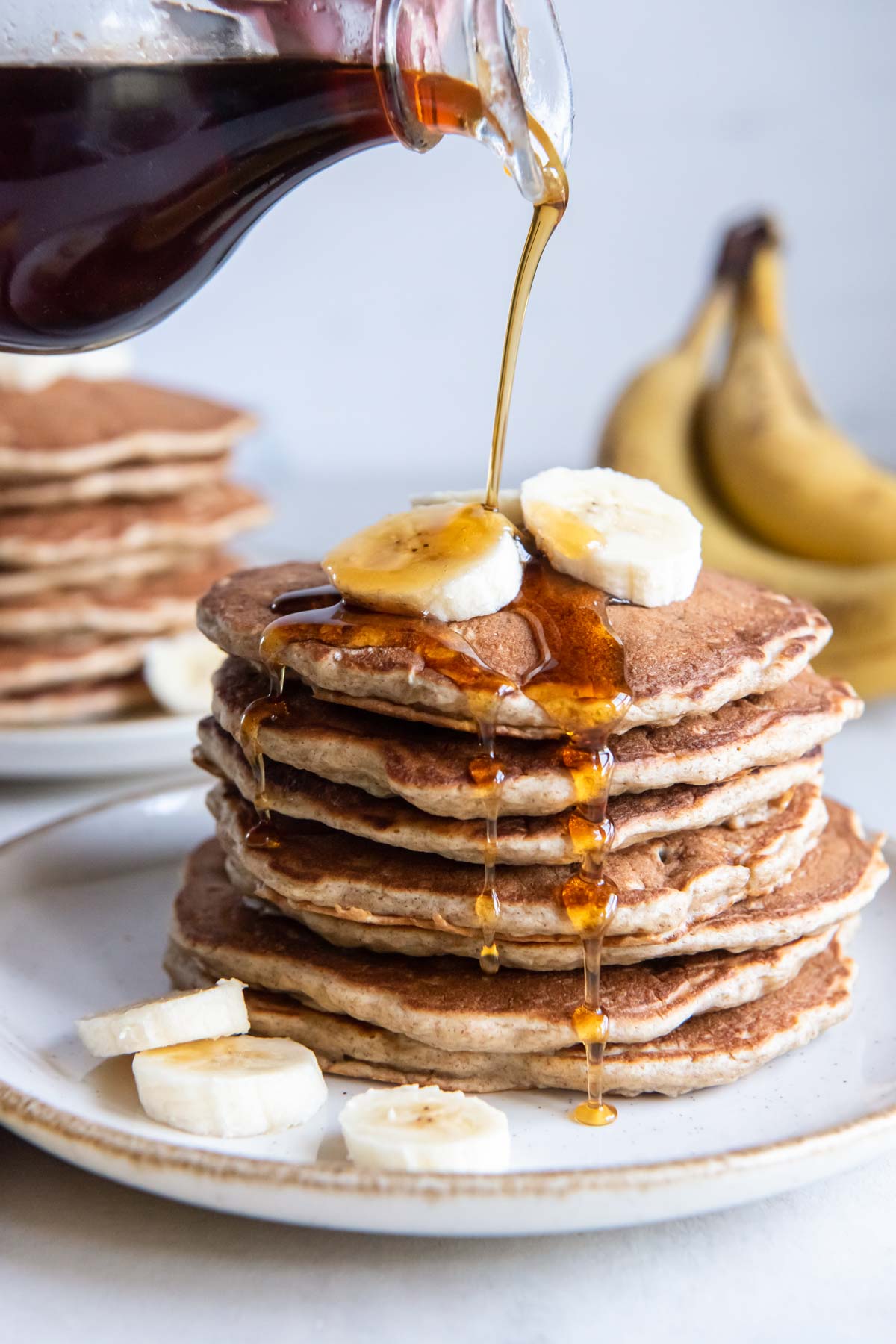 Pouring maple syrup onto stack of 7 banana pancakes topped with fresh banana slices.