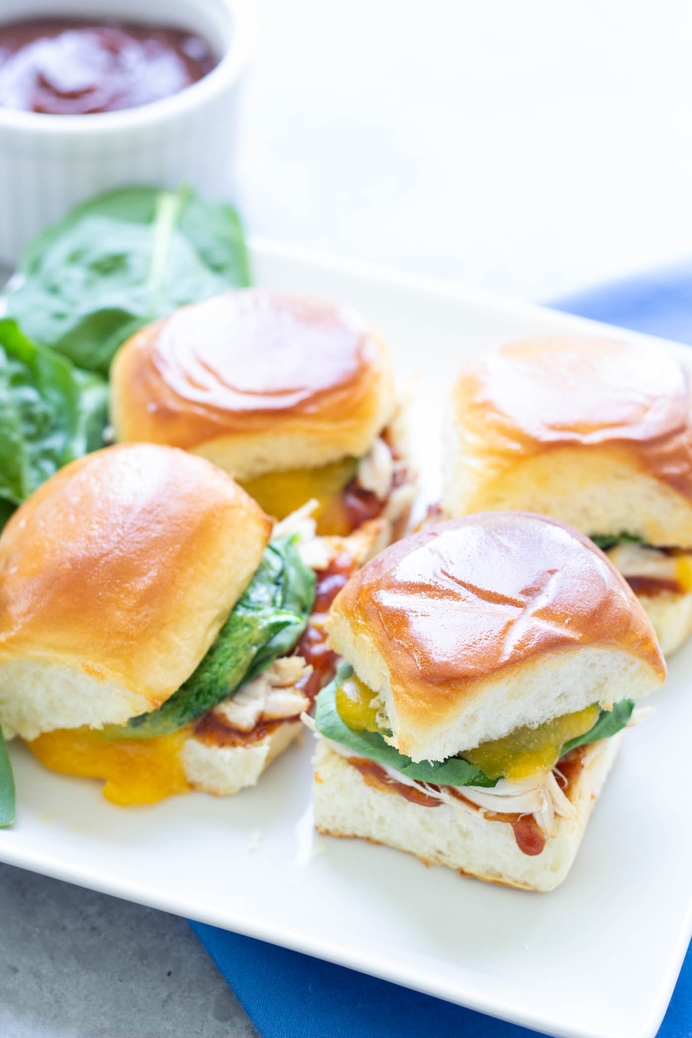 Four BBQ chicken sliders with spinach and cheddar cheese on a plate.