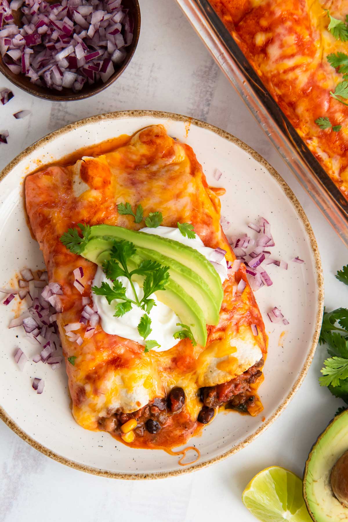 Two enchiladas on a plate, topped with sour cream, avocado, cilantro and red onion.