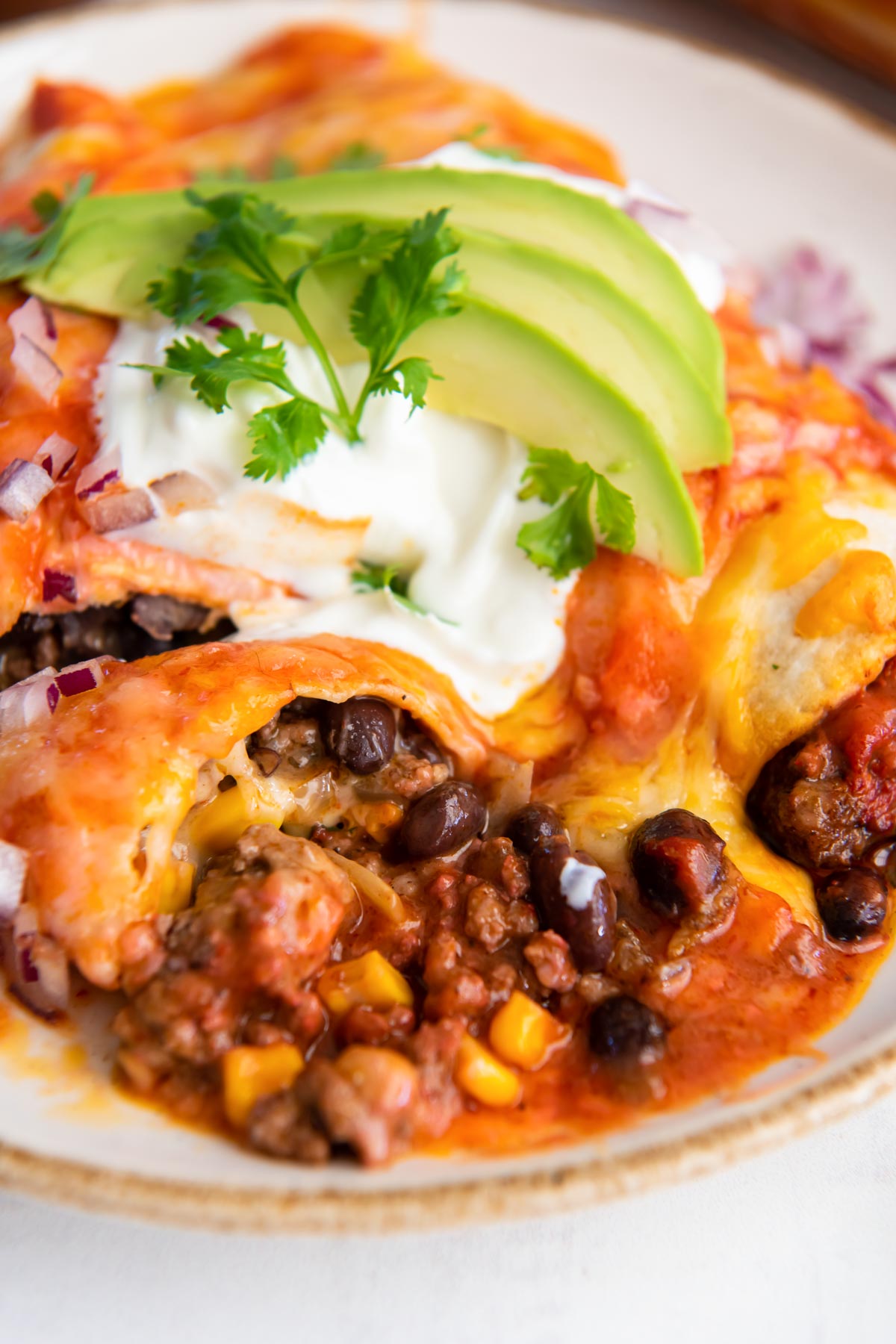 Close up of two beef enchiladas served on a plate with sour cream, avocado and cilantro.