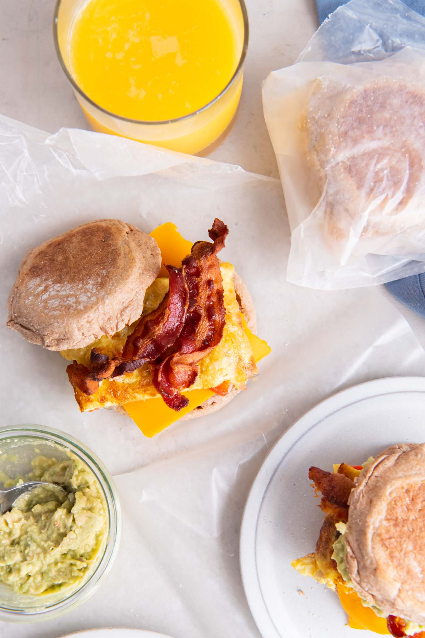 Breakfast sandwich with bacon, unwrapped and wrapped in waxed paper with glass of orange juice nearby.