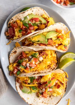Four breakfast tacos with eggs, cheese, bacon, avocado and pico de gallo on a plate.