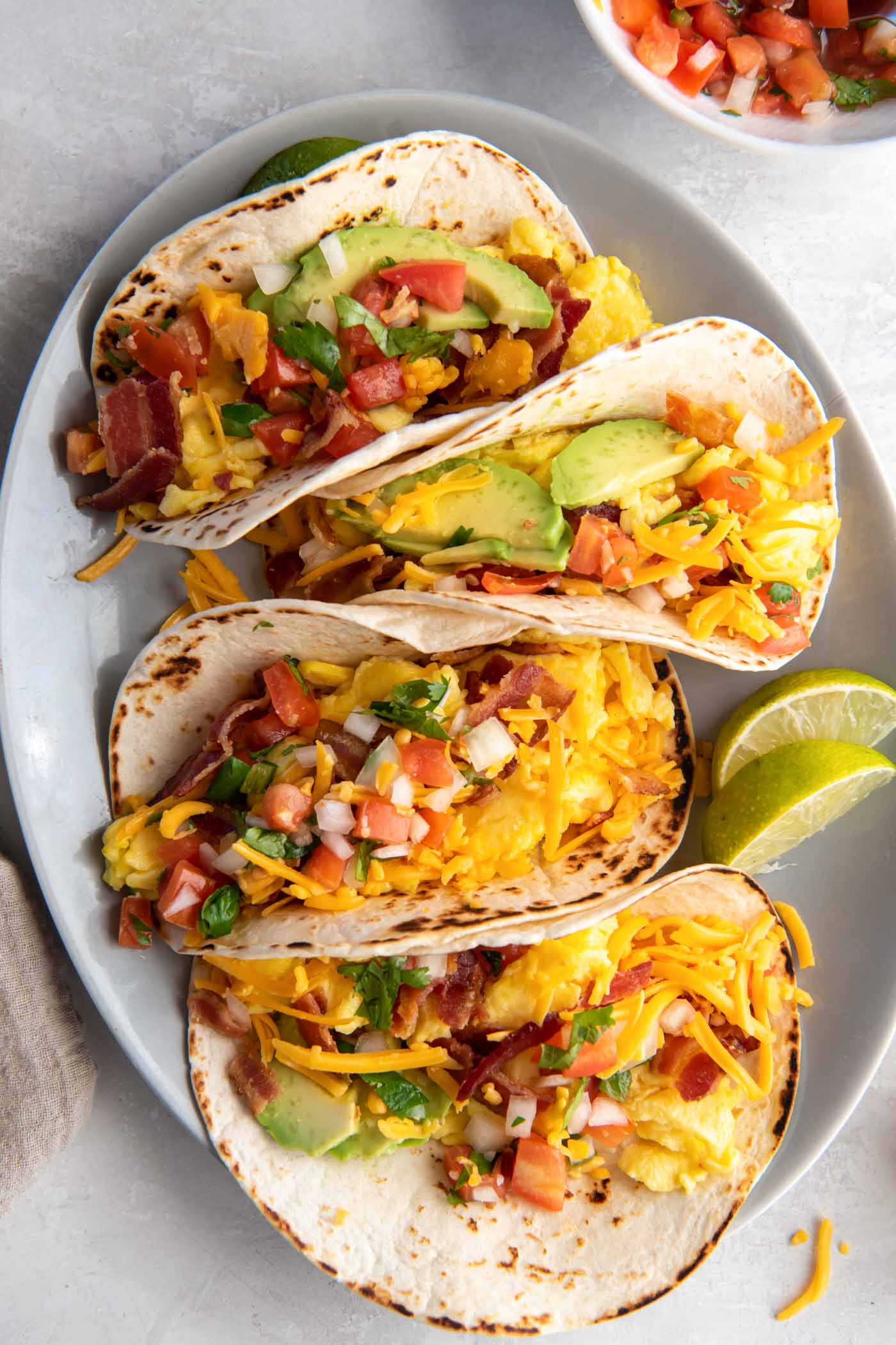 Four breakfast tacos with eggs, cheese, bacon, avocado and pico de gallo on a plate.