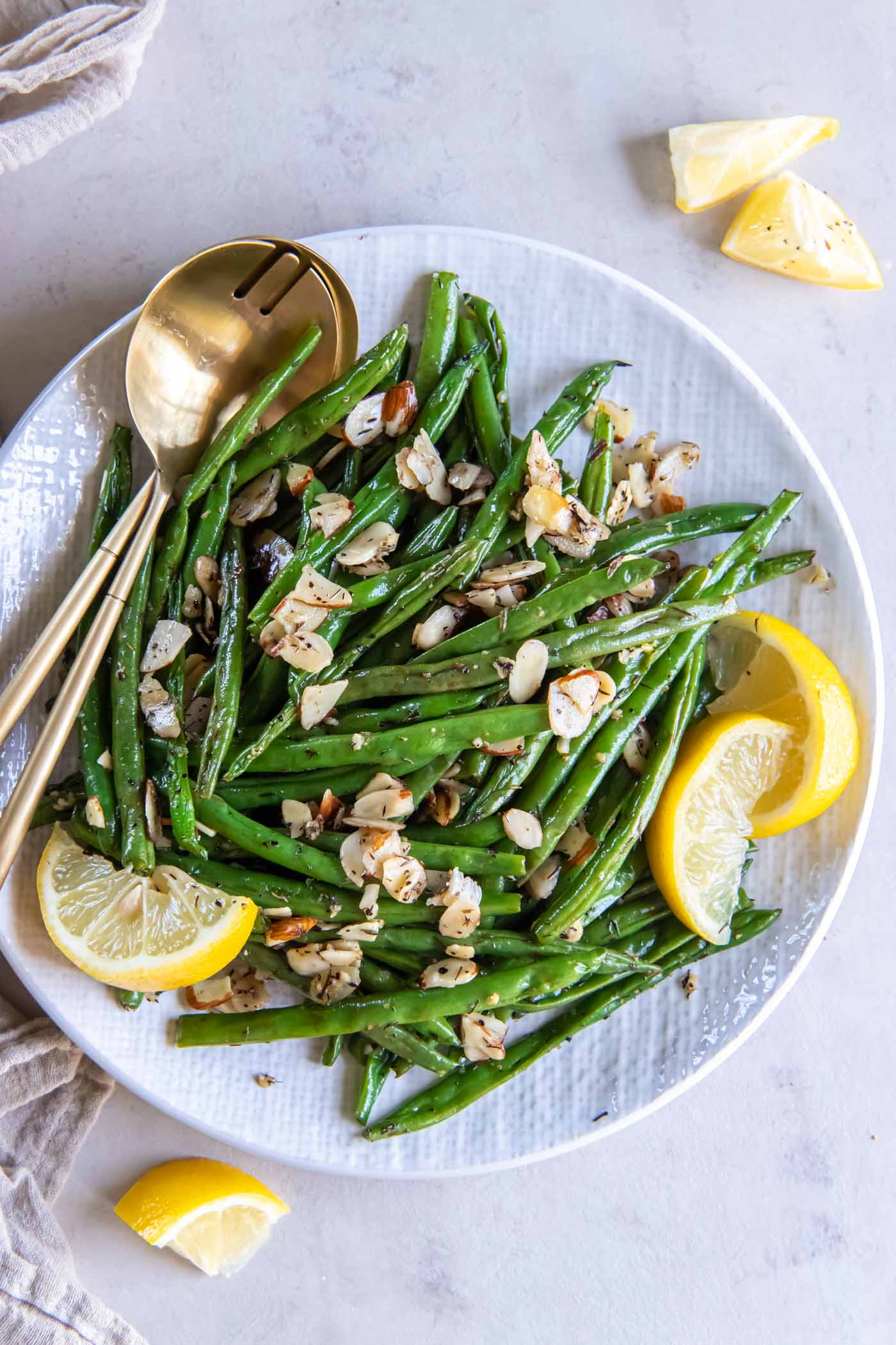Roasted green beans in a serving bowl garnished with lemon wedges, with gold serving spoons.