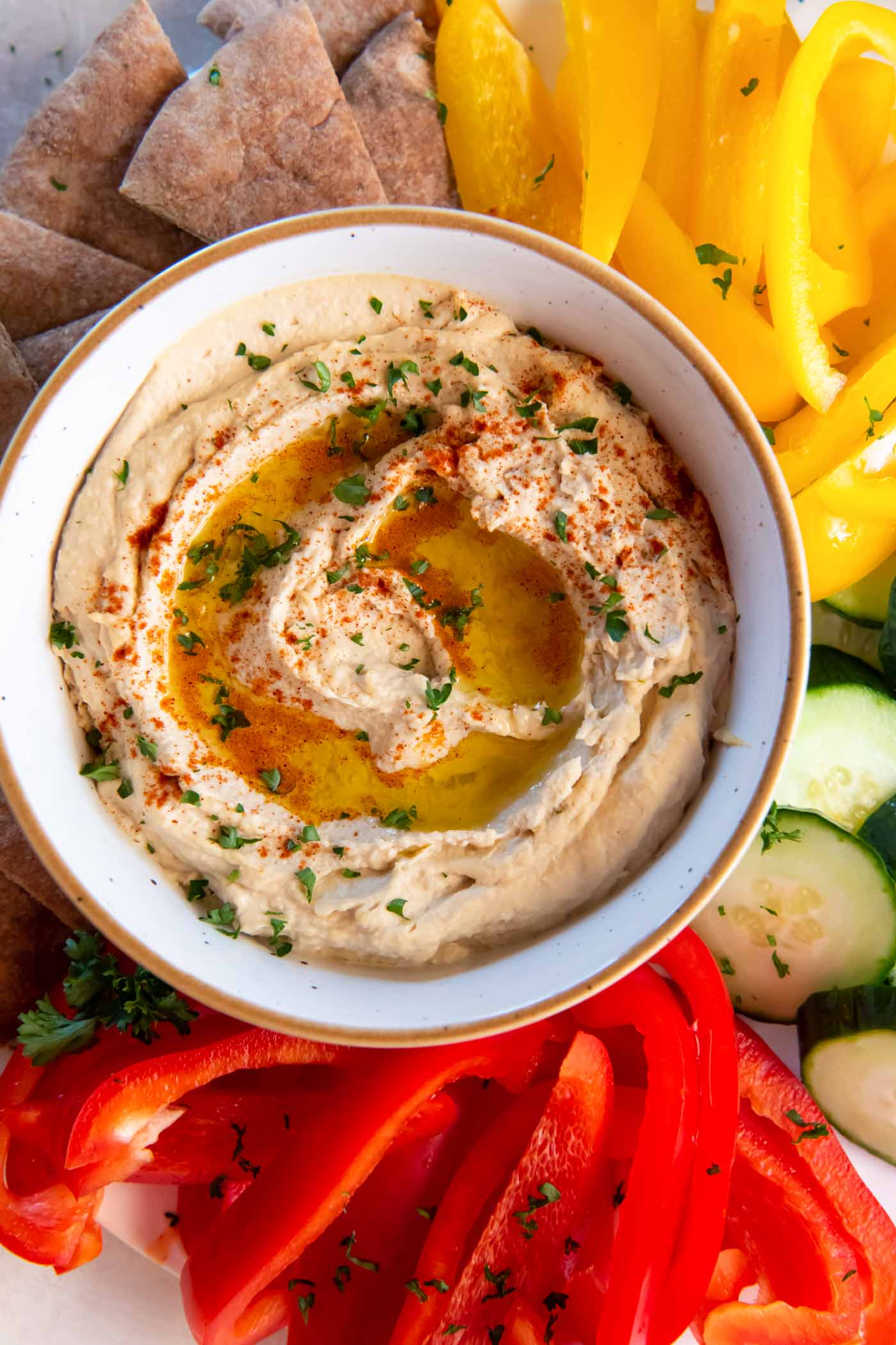 Hummus served in a bowl with sliced bell peppers, cucumber and pita bread around the bowl.