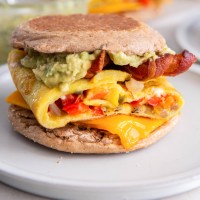 Breakfast sandwich with eggs, peppers, onions, cheddar cheese, bacon and guacamole on an English muffin.