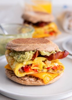 Breakfast sandwich with eggs, peppers, onions, cheddar cheese, bacon and guacamole on english muffin.