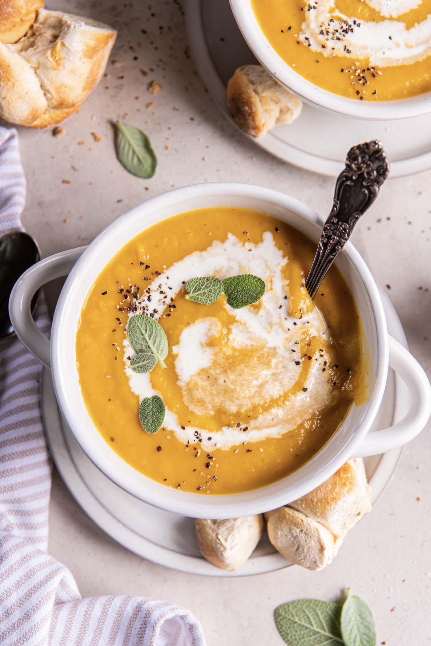 Butternut squash soup served in a soup bowl with a drizzle of cream, a sprinkle of black pepper and fresh sage leaves.