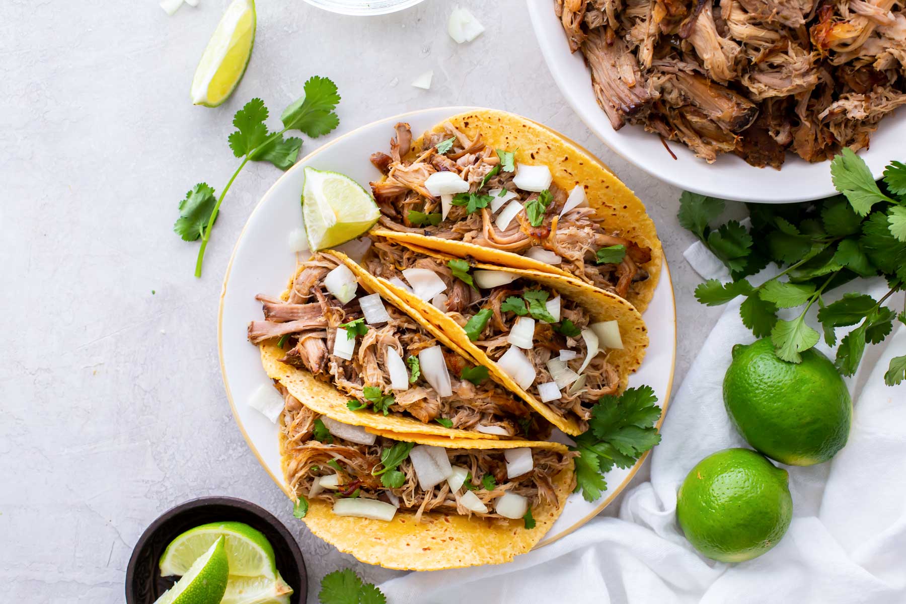 Four carnitas tacos on a plate with bowl of carnitas in background.