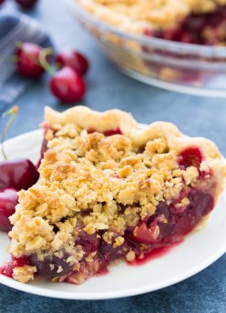 Slice of cherry pie with crumble topping on a white plate, with cherries and a whole pie in the background.