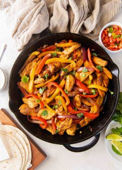 Chicken fajitas in a cast iron skillet with tortillas and fajita toppings around the skillet.