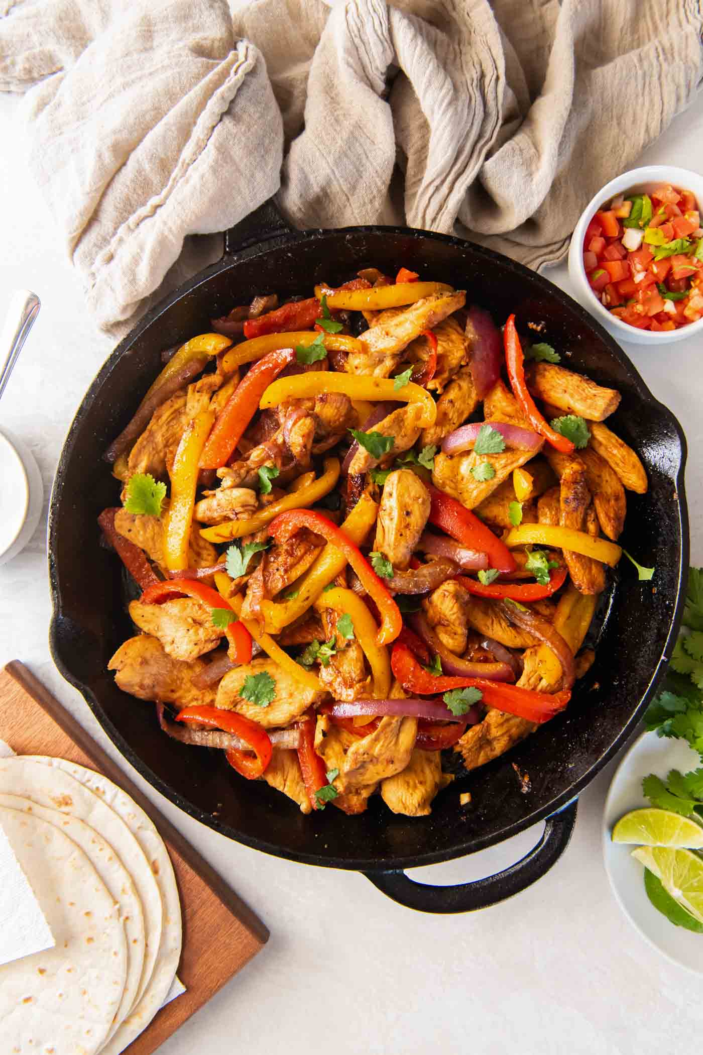 Chicken fajitas in a cast iron skillet with tortillas and fajita toppings around the skillet.