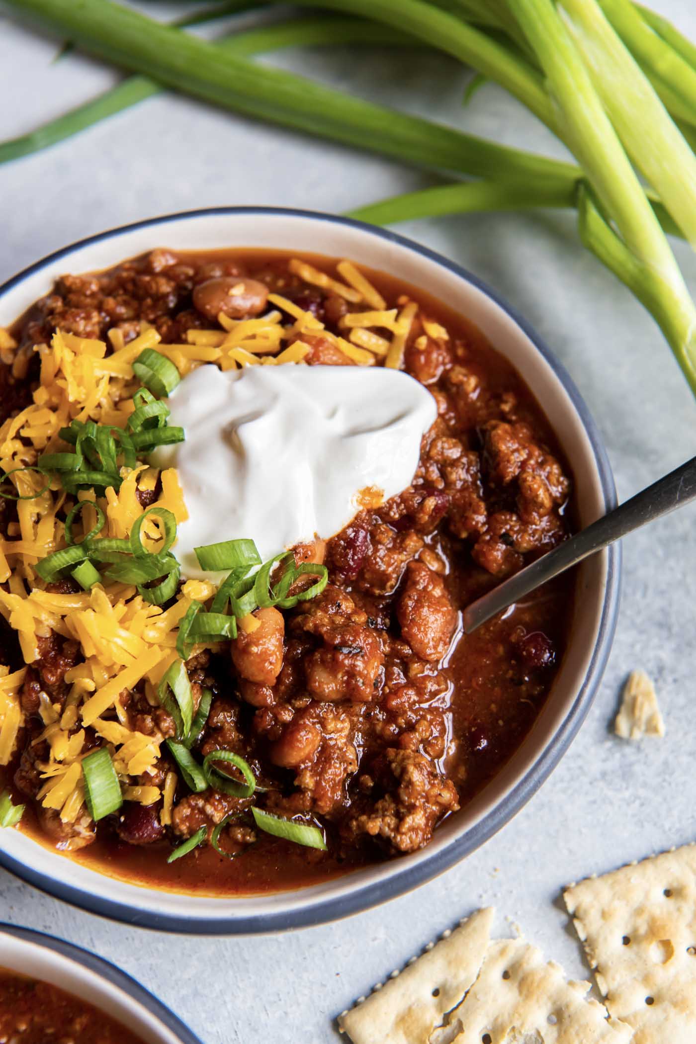 Bowl of chili topped with cheddar cheese, sour cream and green onions with a spoon in the bowl.