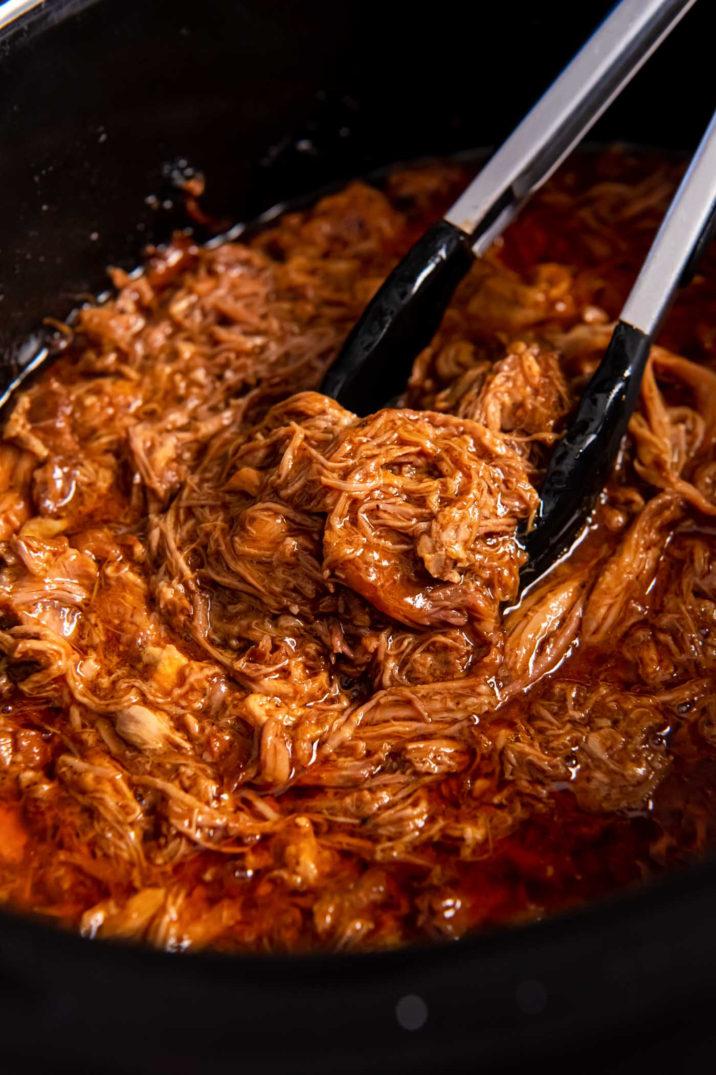 Shredded bbq pulled pork in slow cooker with tongs.