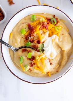 Bowl of crockpot potato soup topped with cheese, sour cream, green onions and bacon with a spoon.