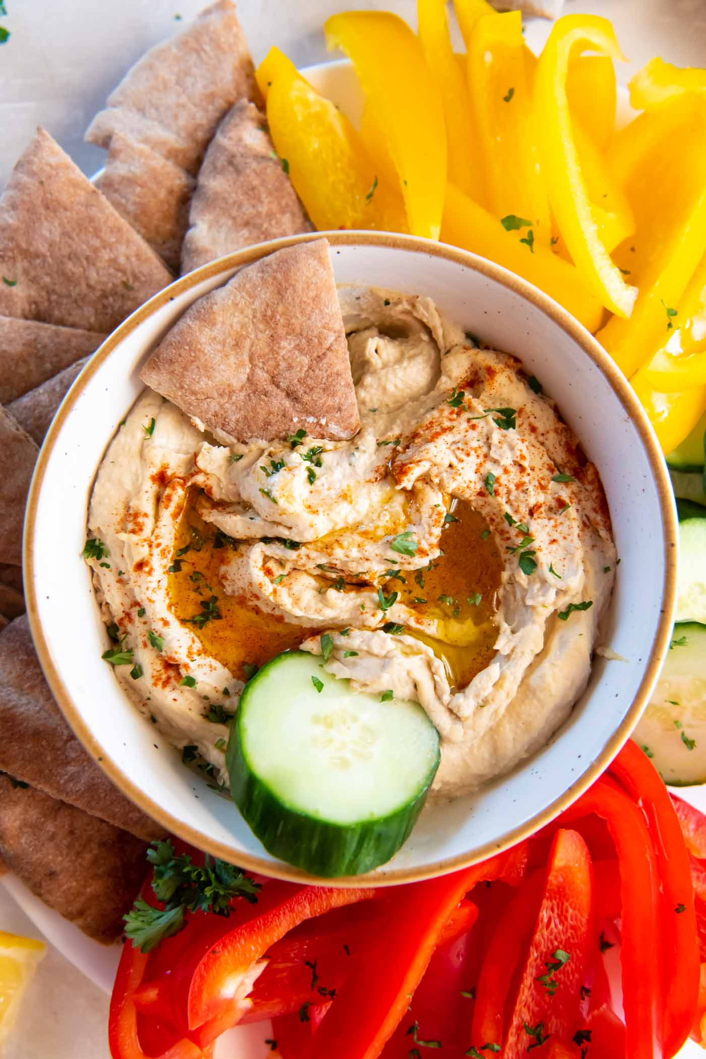 Bowl of hummus with pita bread triangle and cucumber slice in it, with cut vegetables and pita bread around the bowl.
