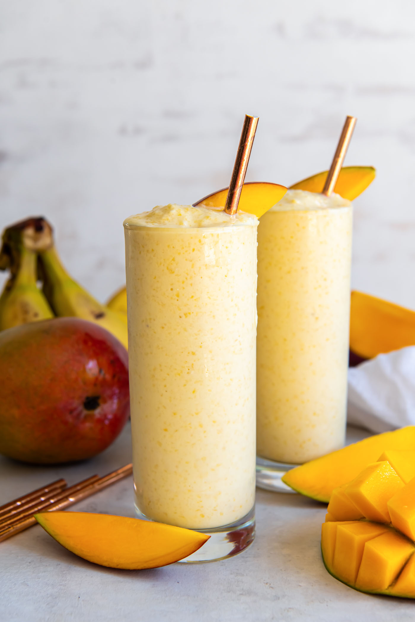 Mango smoothie served in two tall glasses with mango slices and straws.
