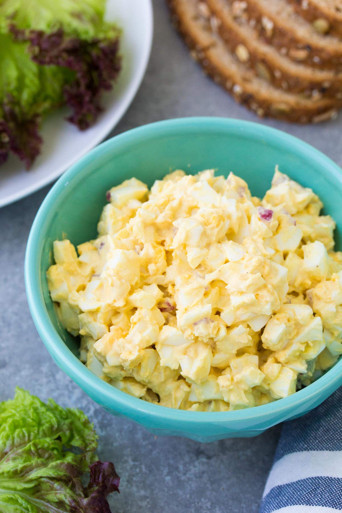 Egg salad in a bowl with lettuce and bread slices in the background.
