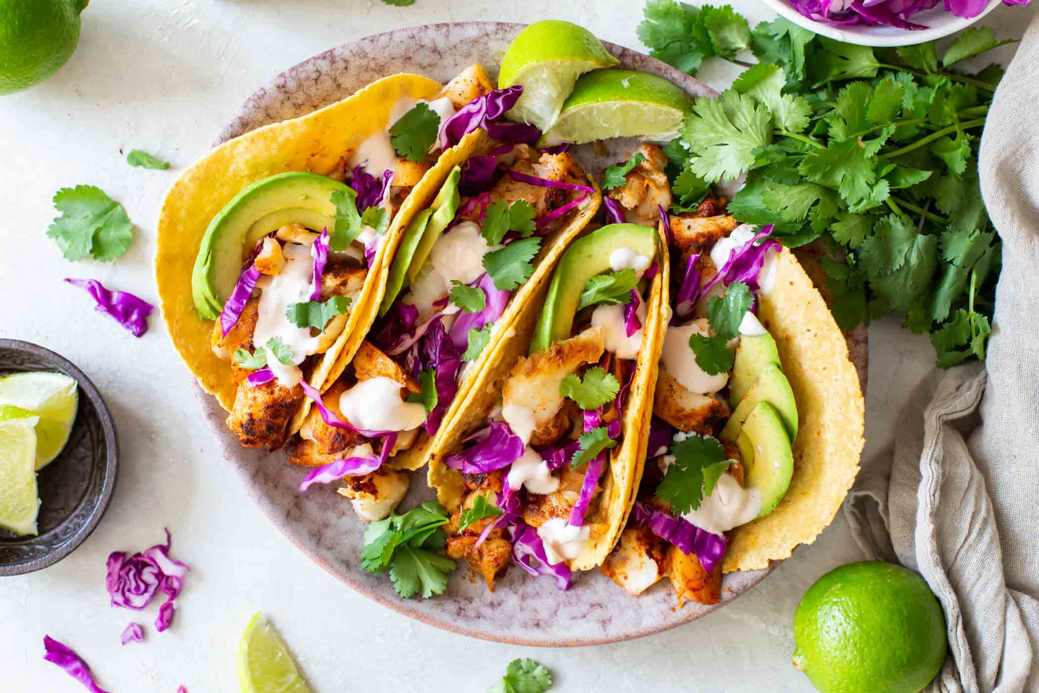 Four fish tacos with fish taco sauce, avocado and cabbage on a plate.