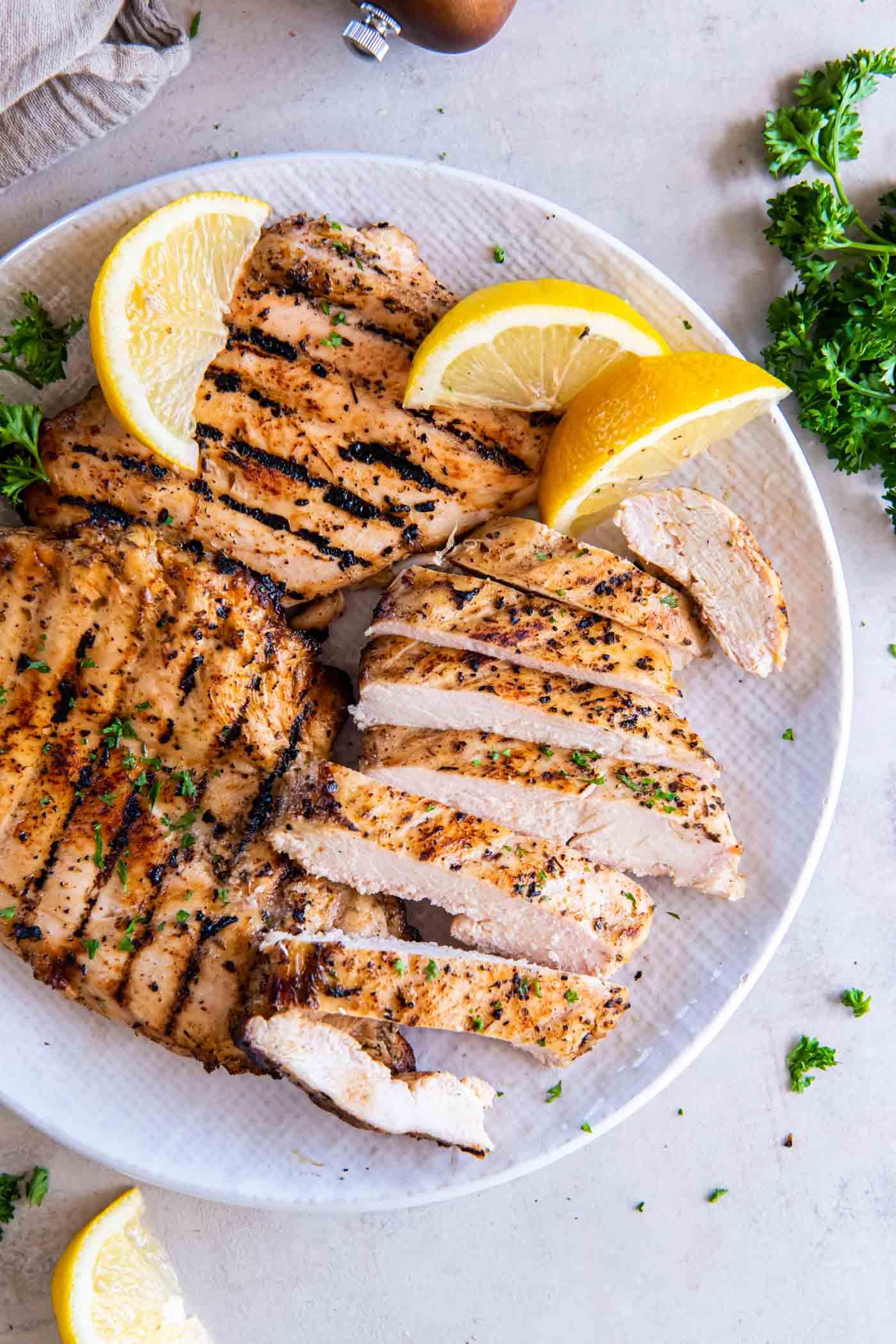 Grilled chicken breasts on a plate with one breast sliced and lemon wedges for garnish.