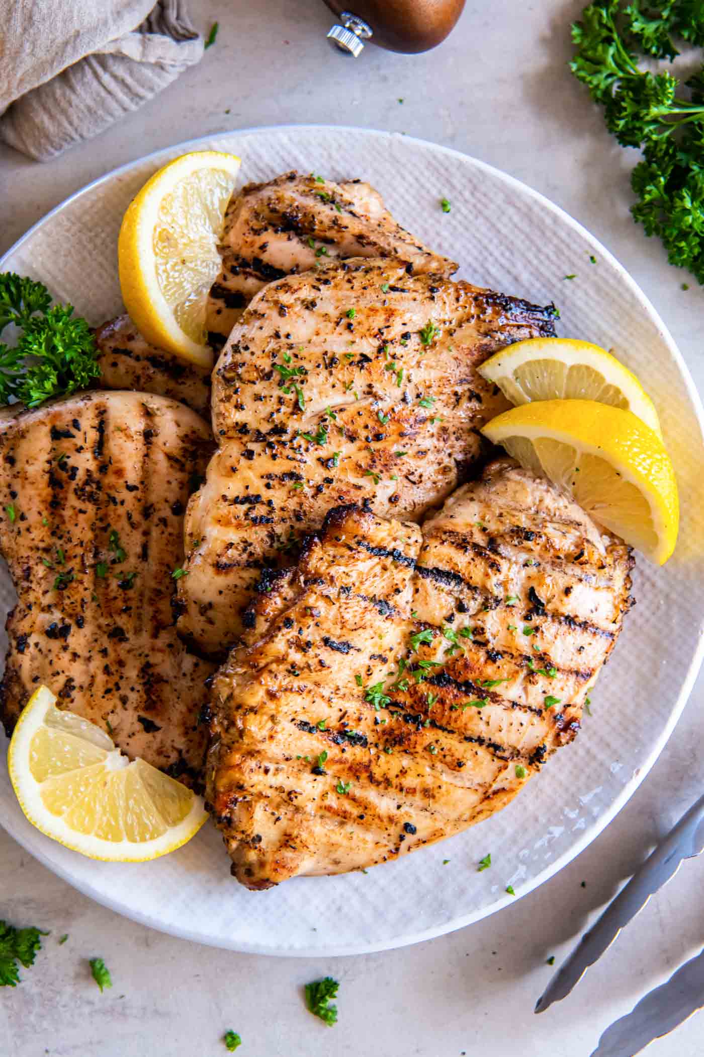 Four grilled chicken breasts on a plate, garnished with lemon wedges and fresh parsley.