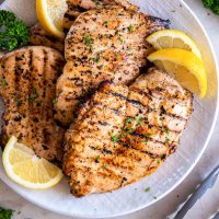 Four grilled chicken breasts on a plate, garnished with lemon wedges and fresh parsley.
