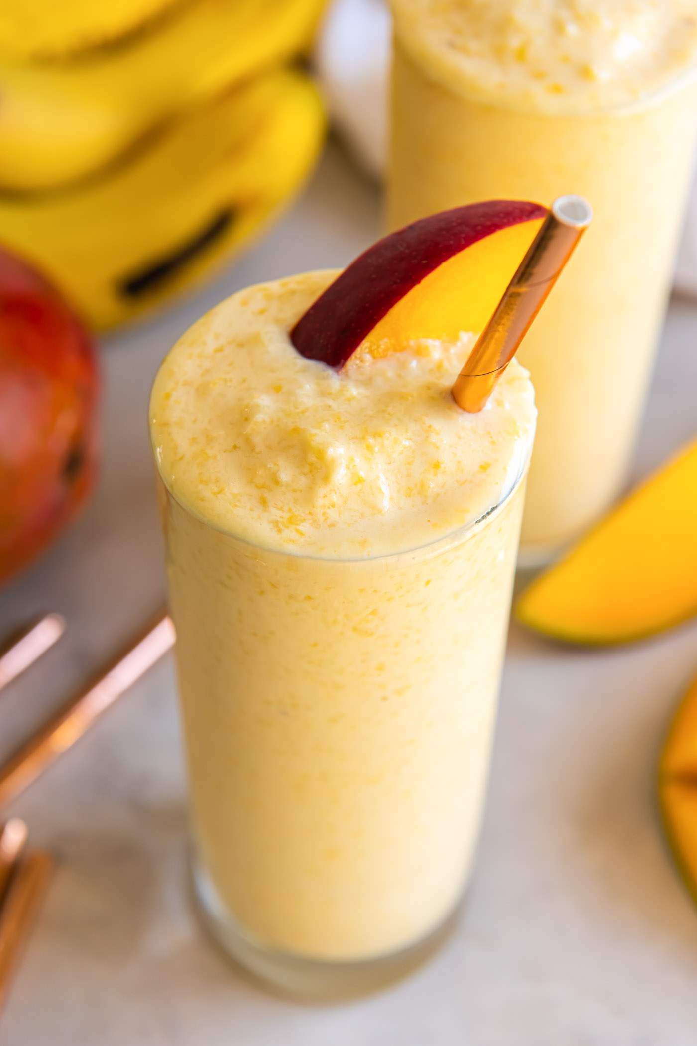 Mango smoothie in a glass with a mango slice and a gold straw.