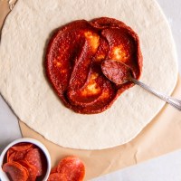 Pizza sauce spread on rolled out pizza dough with a spoon.
