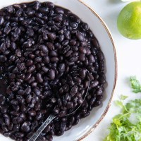 Black beans in serving bowl with spoon, with limes and cilantro on the side.
