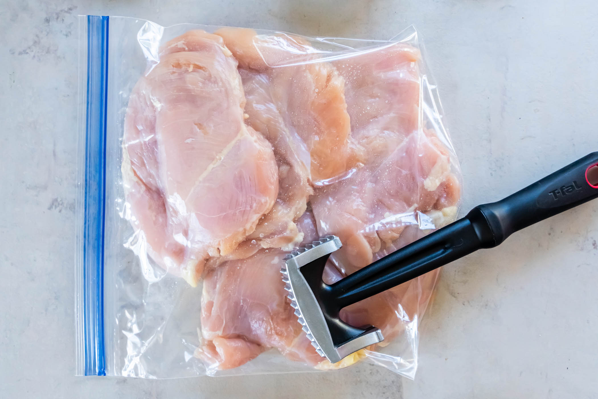 Four raw chicken breasts in zip-top plastic bag with meat mallet resting on bag.