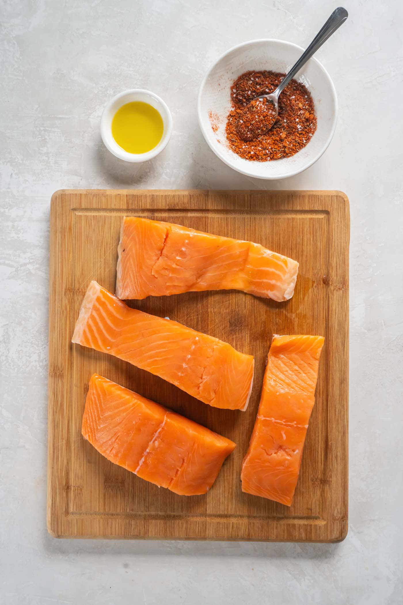 Four raw salmon fillets on a cutting board, alongside a small dish of olive oil and a bowl with salmon seasoning.