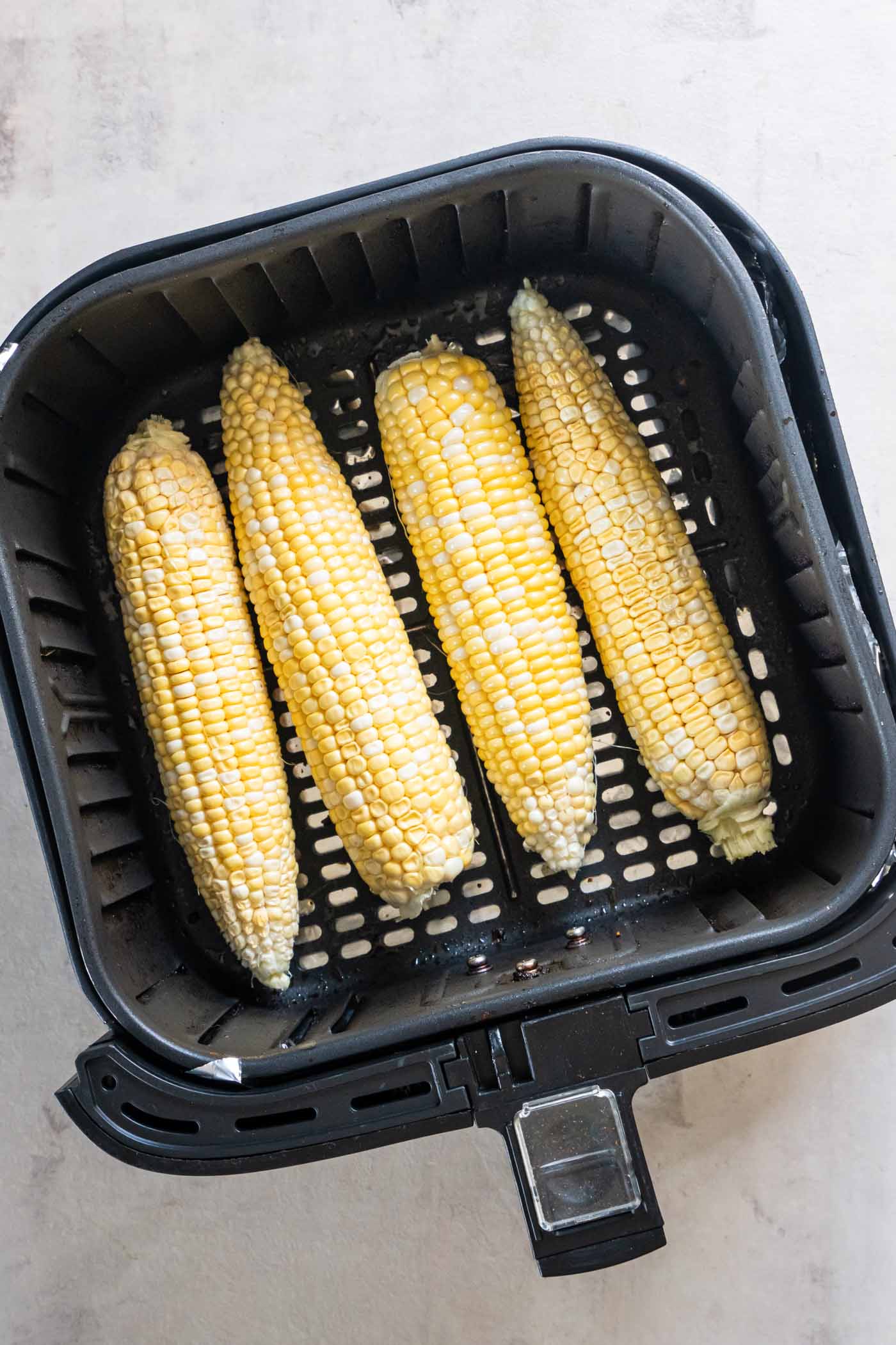 Four uncooked cobs of corn in air fryer basket.