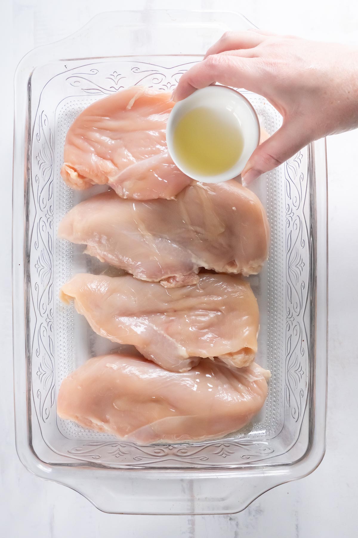 Pouring olive oil over raw chicken breasts in baking dish.