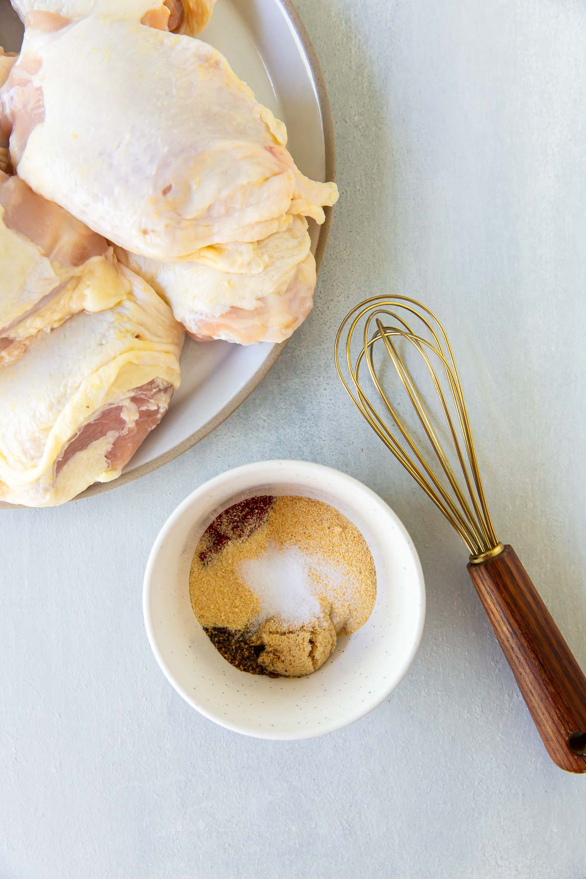 Seasonings in a small white bowl next to a whisk and plate of raw chicken thighs.