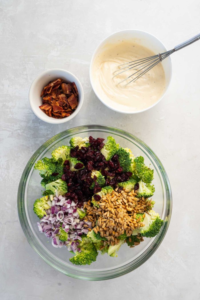 Bowl with broccoli salad dressing, small bowl with crumbled bacon, large bowl with chopped broccoli, red onion, sunflower seeds and dried cranberries.