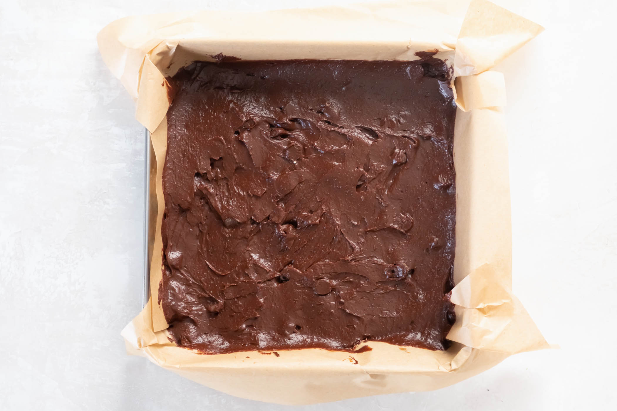 Unbaked brownie batter spread in a metal baking pan lined with parchment paper.