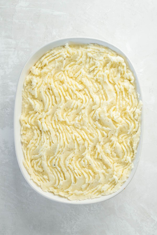 Mashed potatoes added on top of beef mixture in dish, with ridges made on potatoes with a fork.
