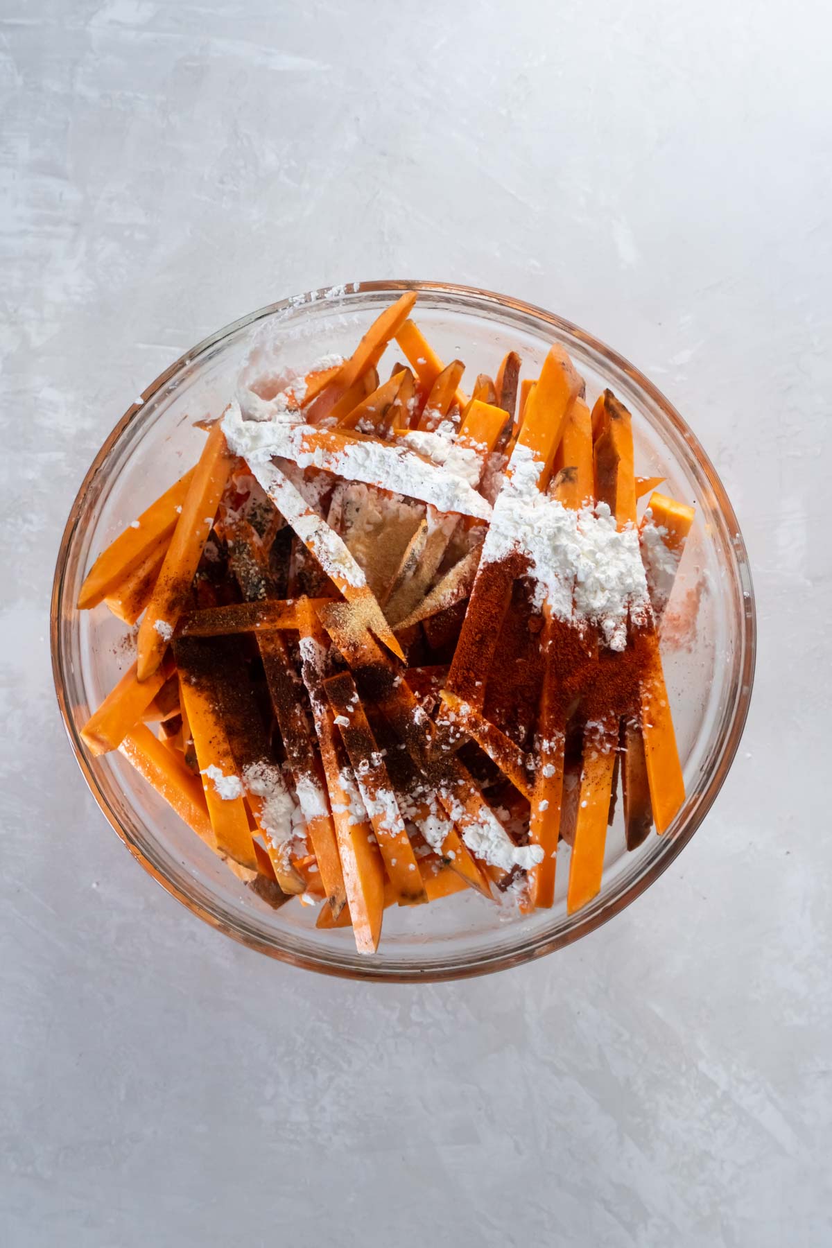 Cornstarch and seasonings sprinkled on cut sweet potato fries in mixing bowl.