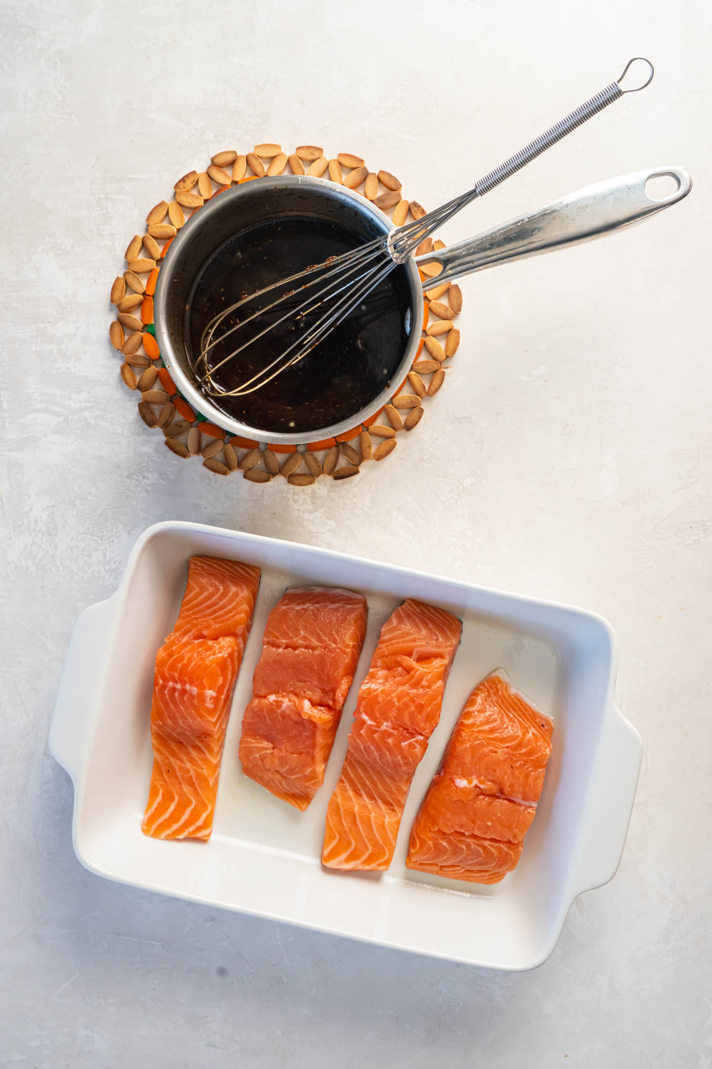 Homemade teriyaki sauce in a small saucepan with a whisk next to baking dish containing four raw salmon fillets.