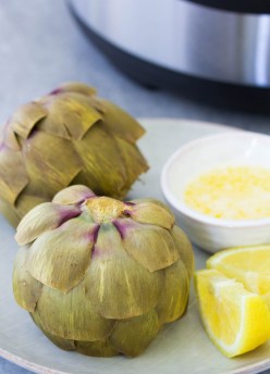 Two Instant Pot artichokes on a plate with lemon and melted butter.