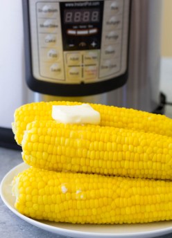 Four ears of corn stacked on a plate with butter on top and Instant Pot in the background.