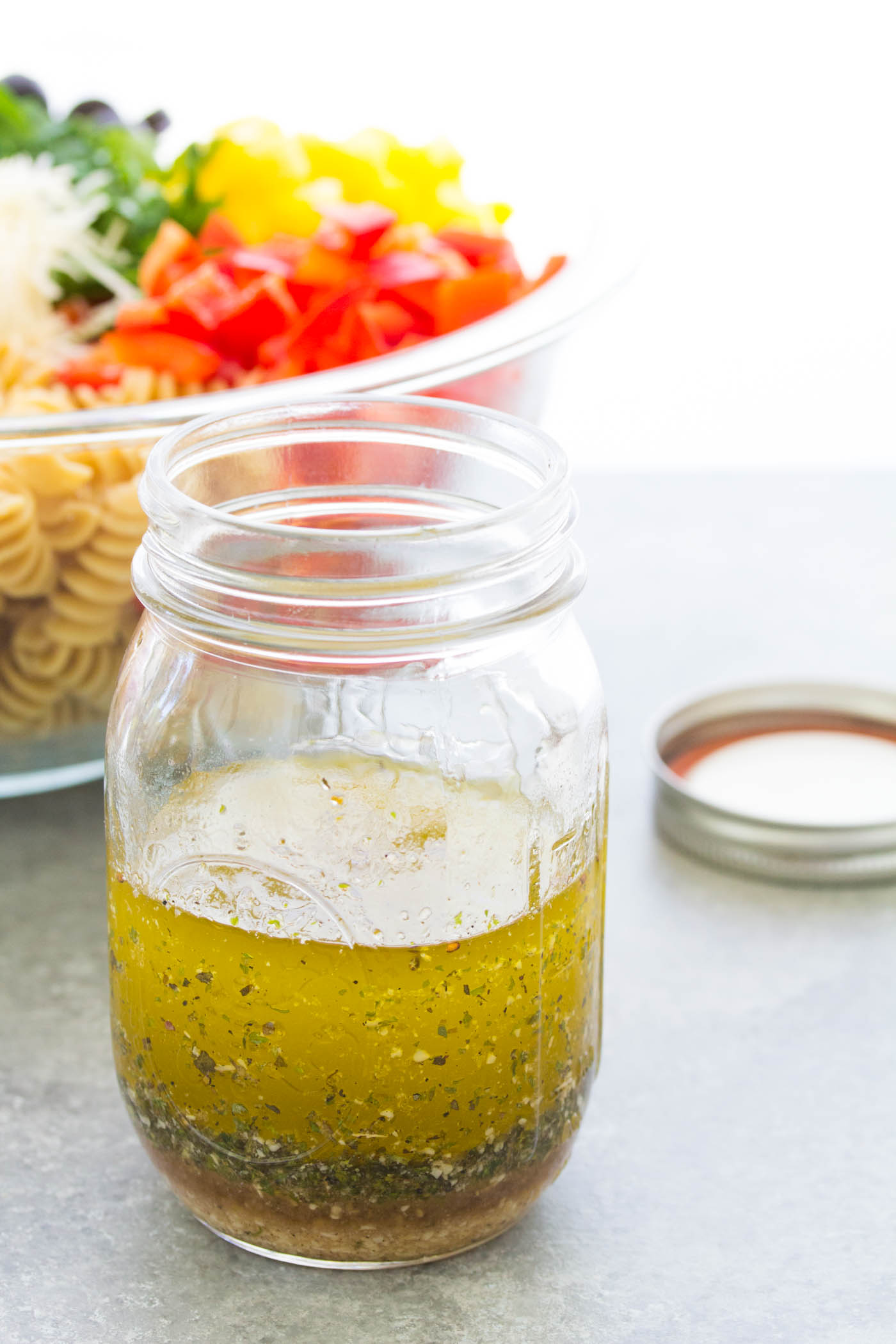 Italian salad dressing in a mason jar with bowl of pasta salad in the background.