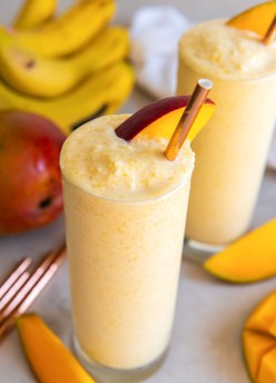Mango smoothie in a tall glass with a mango slice and straw.