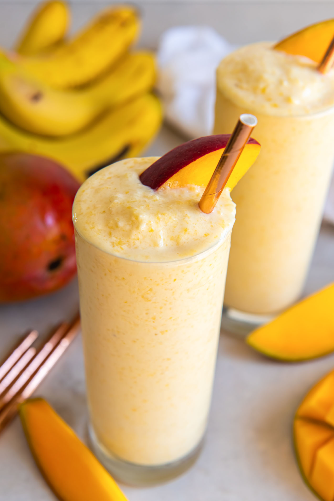 Mango smoothie in a tall glass with a mango slice and straw.