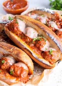 Three meatball sub sandwiches set next to each other with dish of marinara sauce in background.